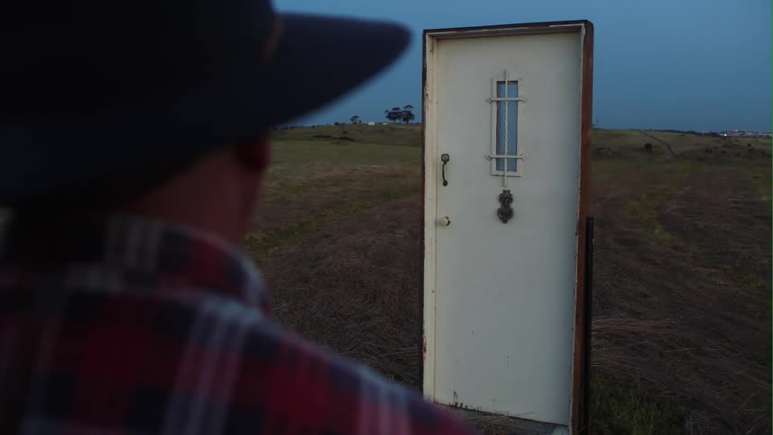 A man wearing a cowboy hat is standing outside in a rural area looking at a door mysteriously standing in front of him