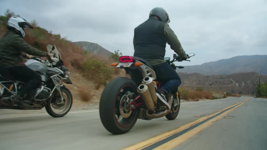 Two people are riding motorcycles on a road with mountains in the distance under a cloudy sky