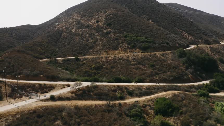 Two motorcyclists drive along curved roads along the coast below rolling hills