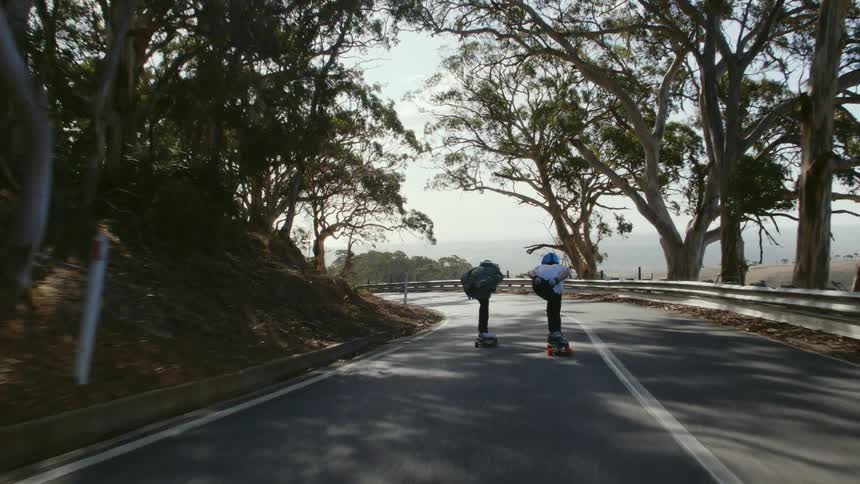 Two people are on skateboards coasting down a hill and they hold hands as the go around a curve overlooking the water
