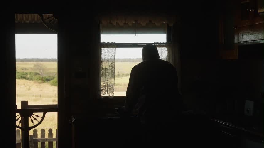A man washes dishes silhouetted by a window on a farm
