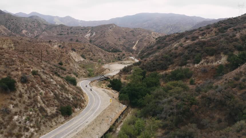 Two motorcyclists ride motorcycles along winding road through scrub covered hills toward misty valley on the horizon