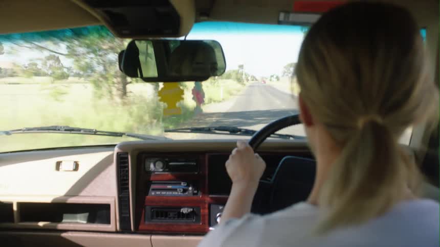 A woman is driving an automobile on a rural road with bread and milk in the seat next to her