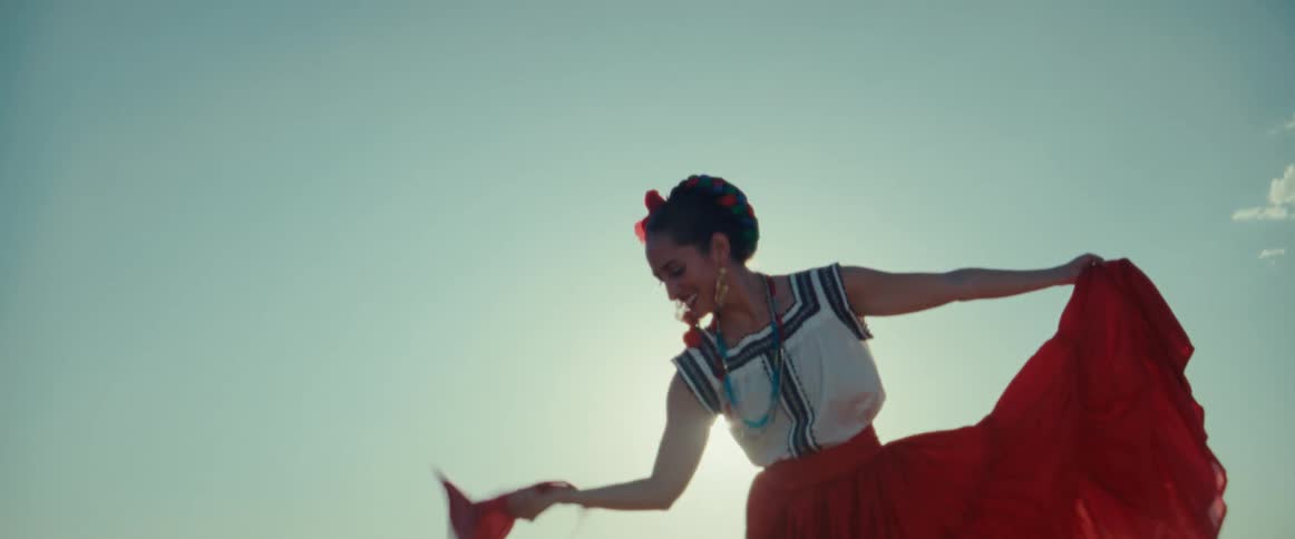 A woman wearing a long red skirt with flowers in her hair and waving a red scarf dances outside as the sun shines brightly upon her