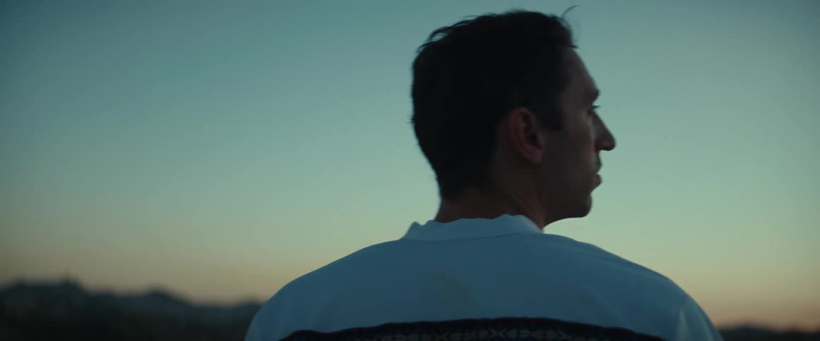 A man stands outside at dusk and puts a hat on