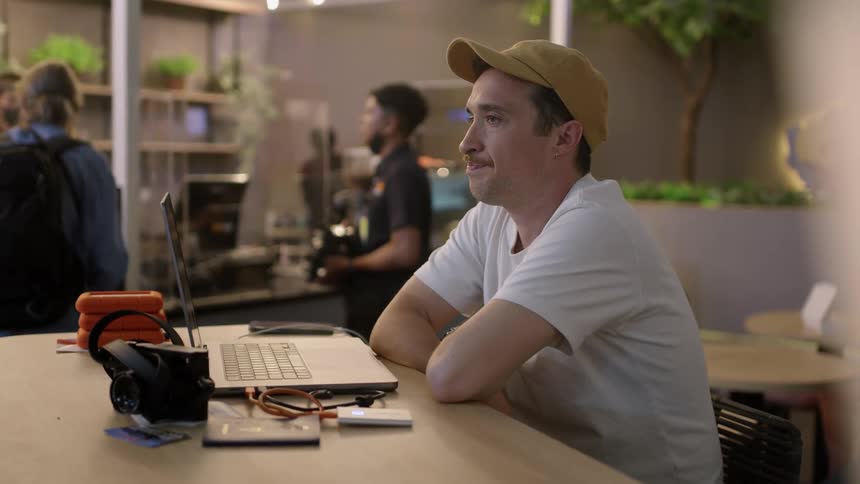Man with yellow cap mustache and white shirt sits at table using laptop at a cafe