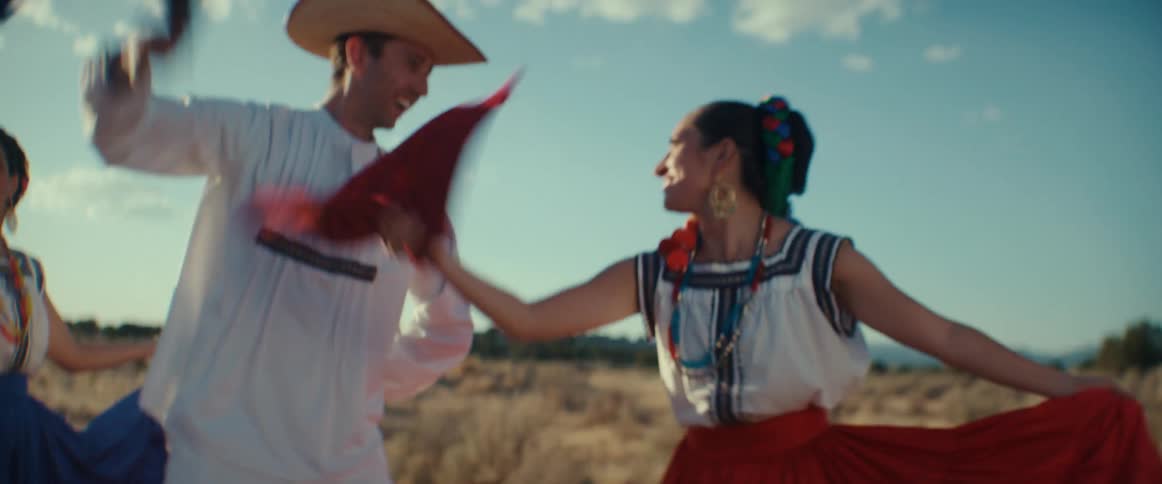 Mexican folk dancers perform group dance in field on sunny day while wearing traditional costumes