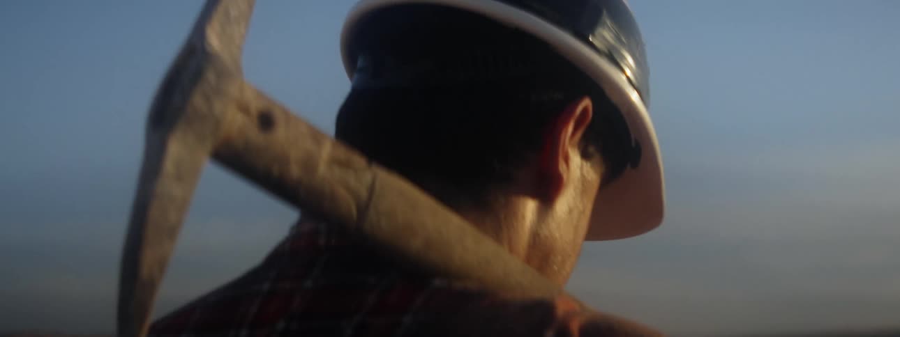 Man with hardhat and plaid shirt carries pickax over shoulder in field