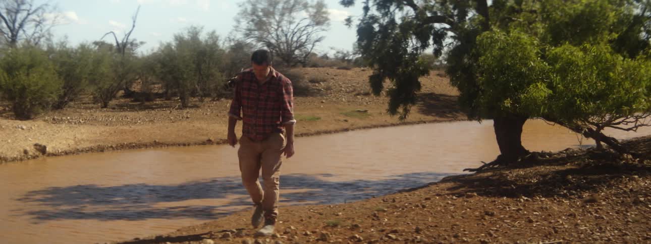 Man with red plaid shirt and brown pants walks on river bank