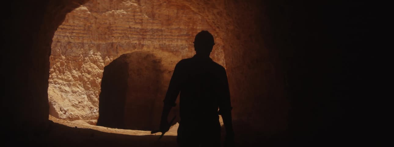 A man wearing a hardhat and carrying a pickax is walking towards a mine shaft
