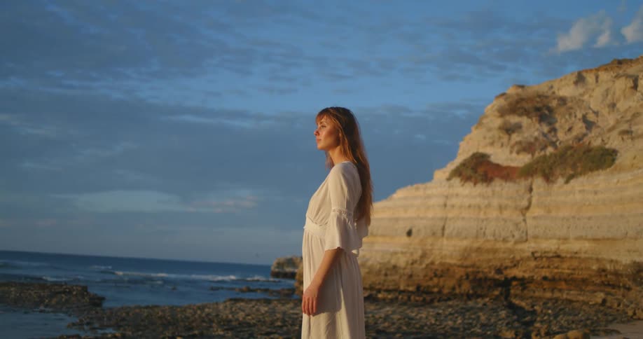A woman in a white dress stands along the ocean shore while looking into the distance