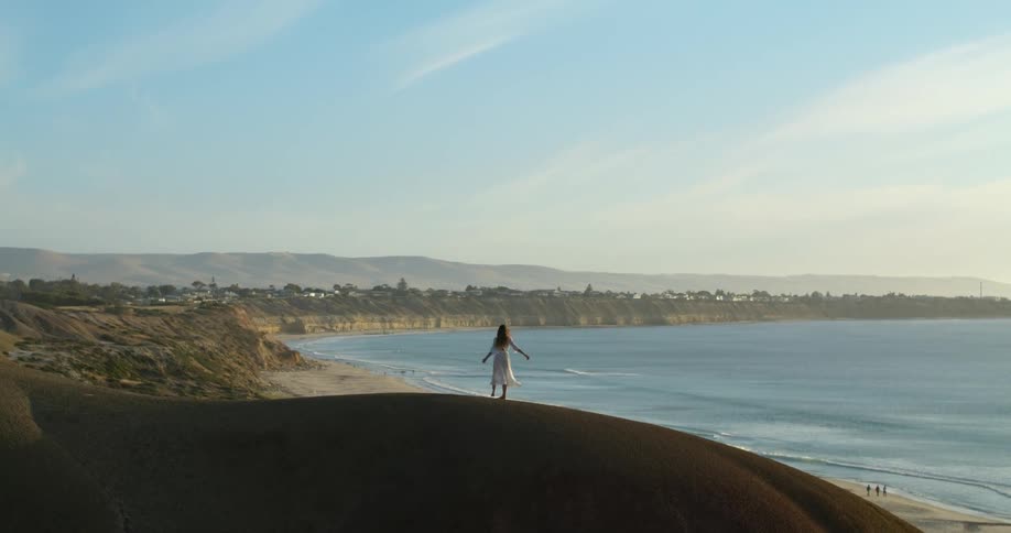 Young woman wearing a white dress spins over a hill next to the ocean at sunset