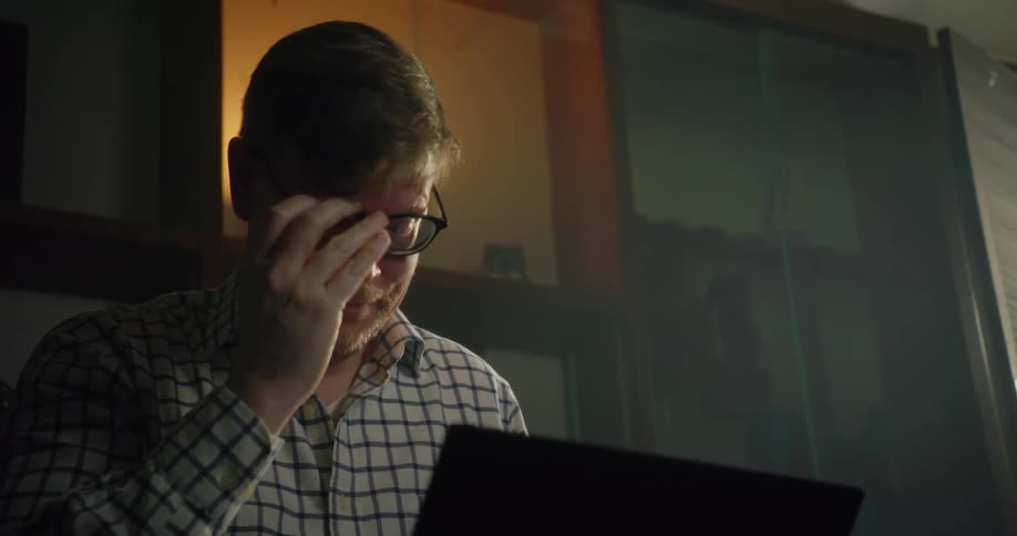 A man looks at his laptop and then takes his glasses off and rubs his eyes and runs his hand through his hair