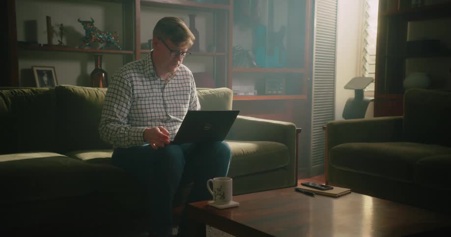 A man sits on a couch at home and types on a laptop that is on his knee