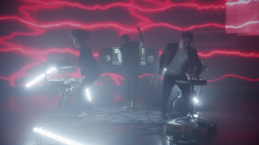 Band members on a small stage jumping and dancing as they play electronic music with a flashing screen behind