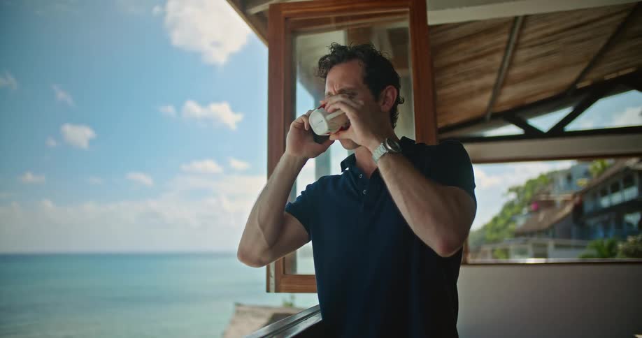 A man talks on his phone while drinking something out of a mug in a house on the beach
