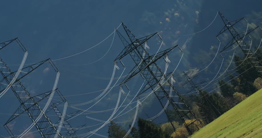 Multiple transmission towers on a green field near pine tree forests close to the mountains