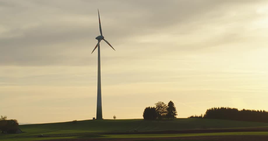 The sun shines through clouds in a hazy sky as a wind turbine spins high above trees