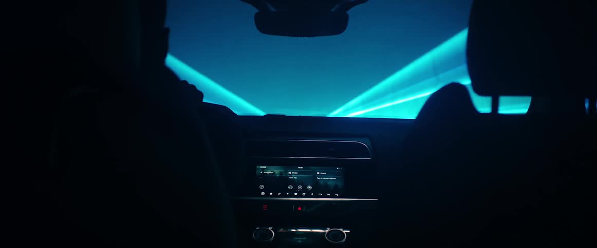 A person sits in the driver seat of an automobile while beams of blue light are projected in front of it