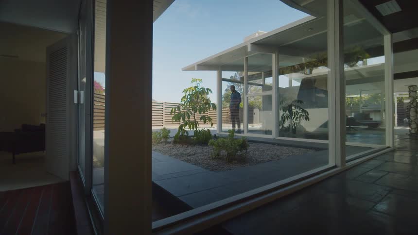 A man looks out the floor to ceiling window of his modern home