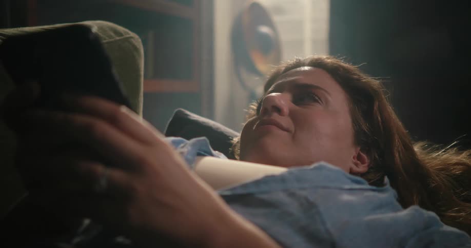 Emotional woman with brown wavy hair white top and blue shirt lies on couch holding smartphone