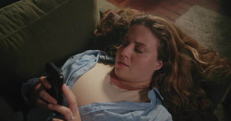 A woman checks her smartphone while lying on a couch in her living room