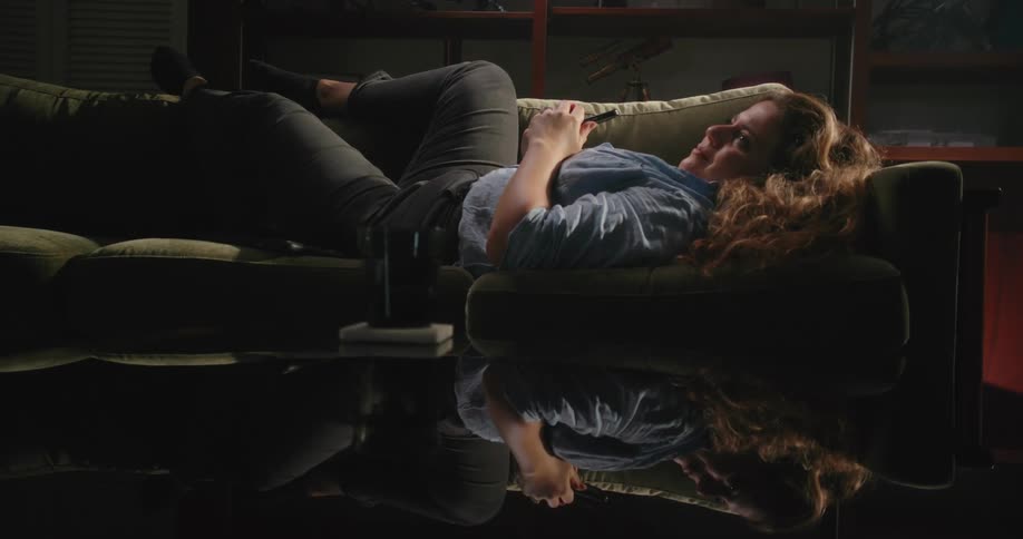 Young happy woman reclined on a couch in the cozy living room smiles while browsing on a smartphone
