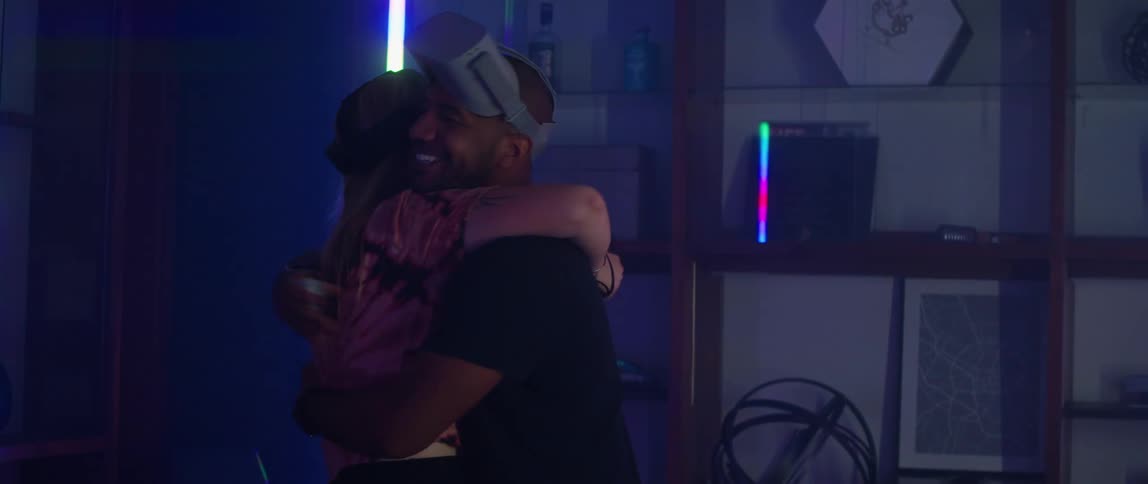 Bald man with black shirt and woman with blonde hair and pink blouse take off virtual reality headsets and hug each other in room