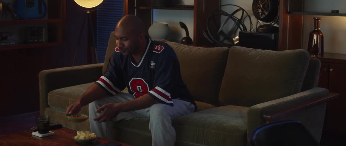 Bald man wearing jersey shirt and gray sweat pants eats snack and watches sports match on television in living room at home