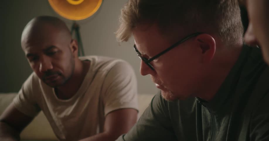 Two men are sitting on a couch looking down at something and talking