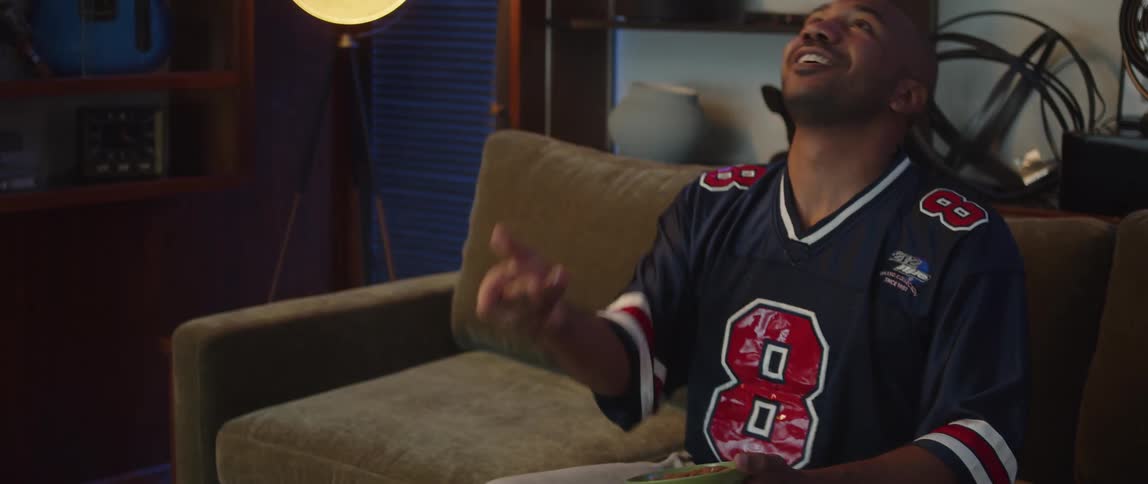 A man wearing a sports jersey is sitting on a couch watching television and holding a bowl of snacks as he cheers and celebrates