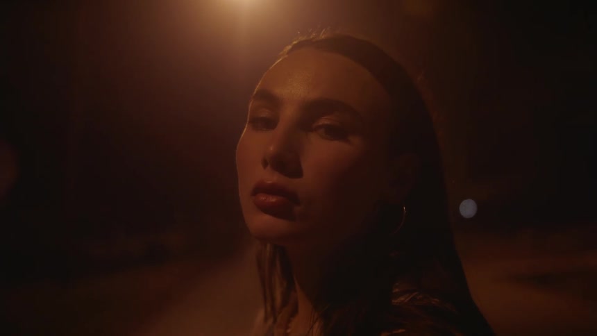A woman is standing in the darkness illuminated with orange light as she moves her hair behind her ears with her hands