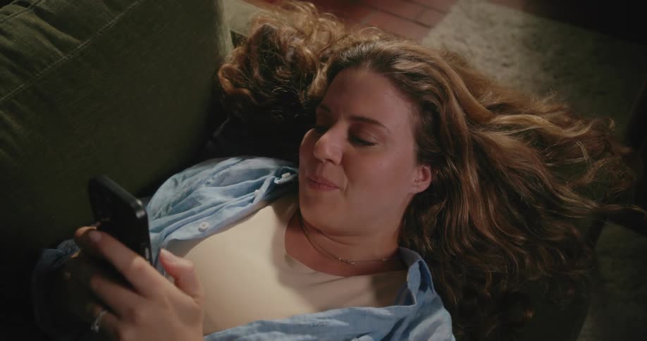 A woman is lying on the living room couch smiling as she texts on her cell phone