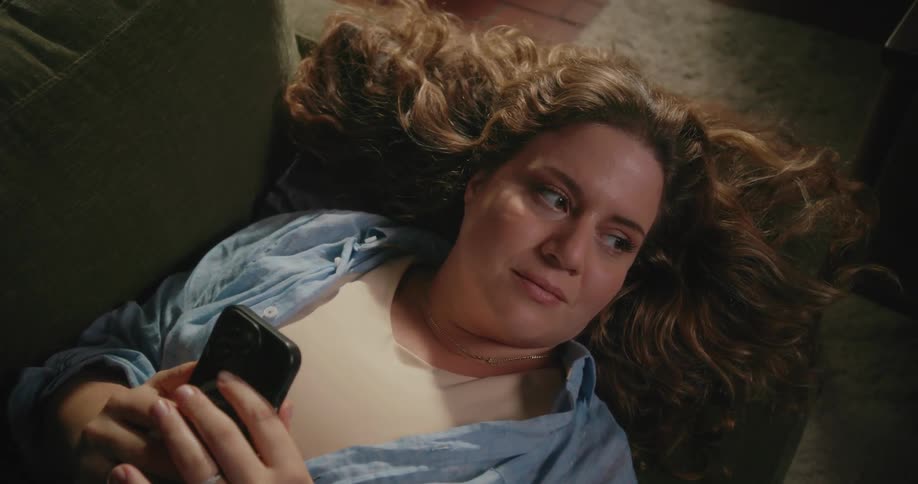 A woman lays on her back on the couch while looking at her smartphone screen