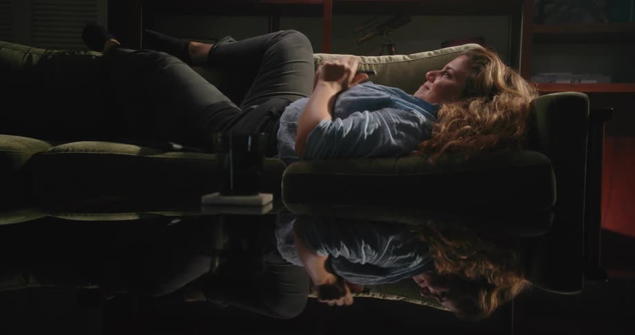 Woman with brown wavy hair blue shirt and black pants lies on couch using smartphone in living room