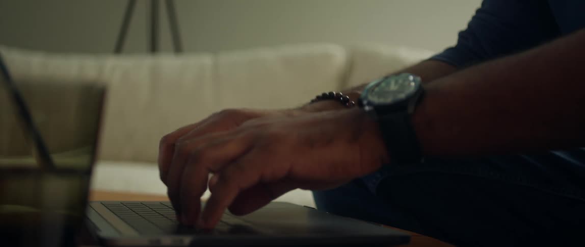 Man with black shirt and wristwatch types on laptop keyboard in living room at home