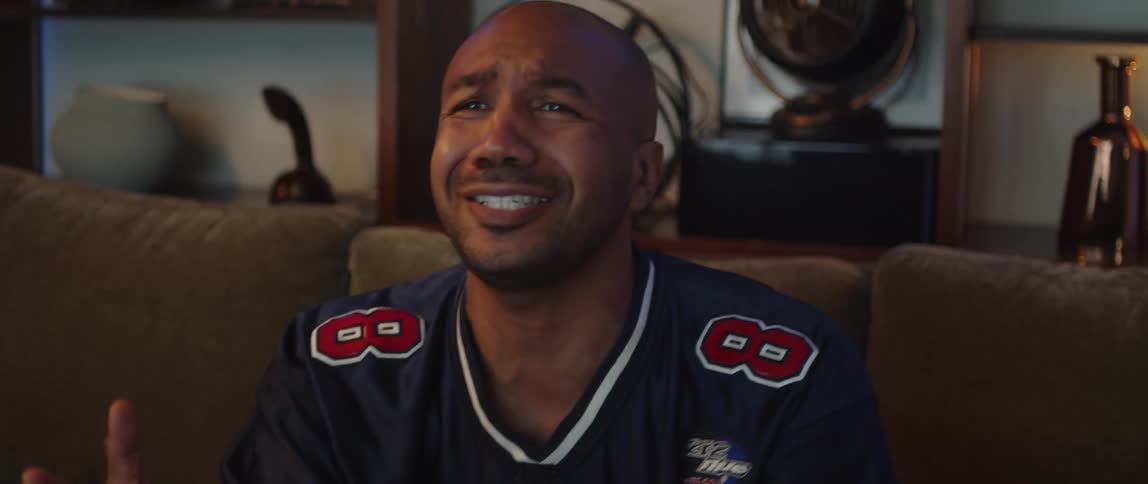 A man wearing a sports jersey is sitting on a couch watching television holding a bowl of snacks and getting emotional