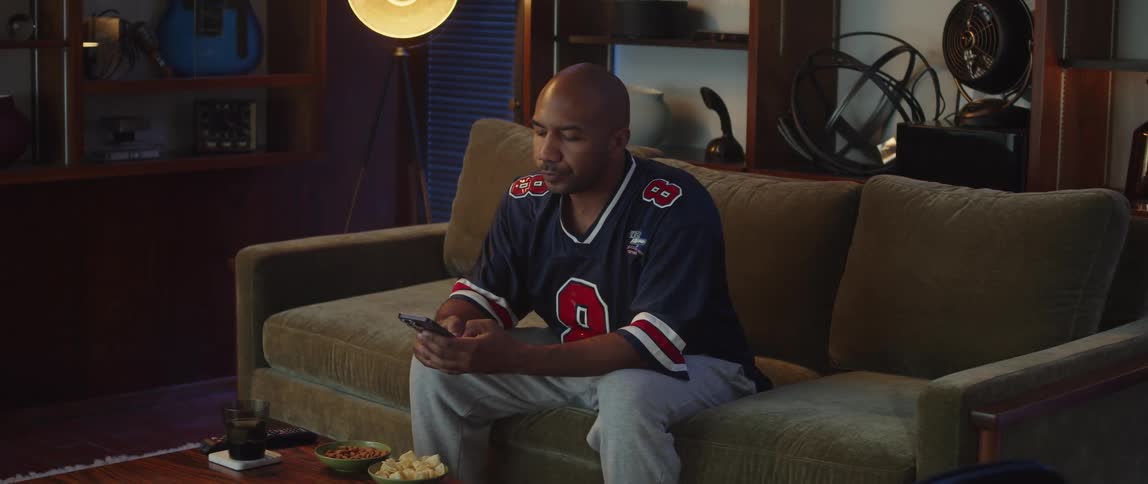 A man eats chips while scrolling through his phone on the couch