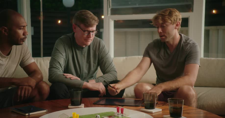 Three men play a board game using a tablet
