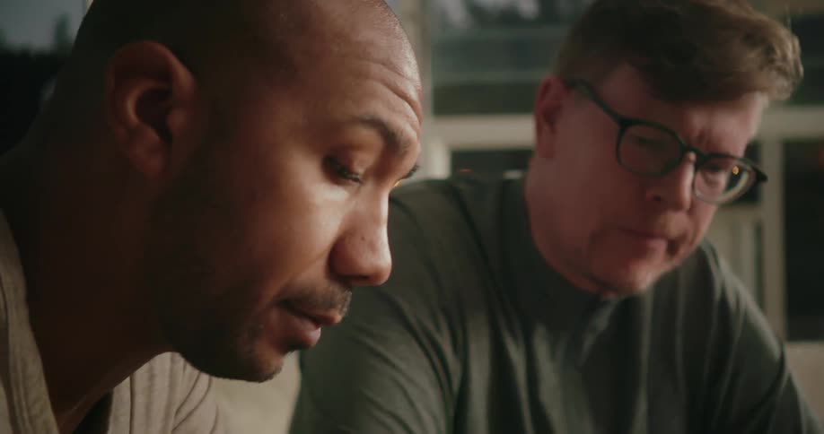 Two men are sitting on a couch looking down at something and talking