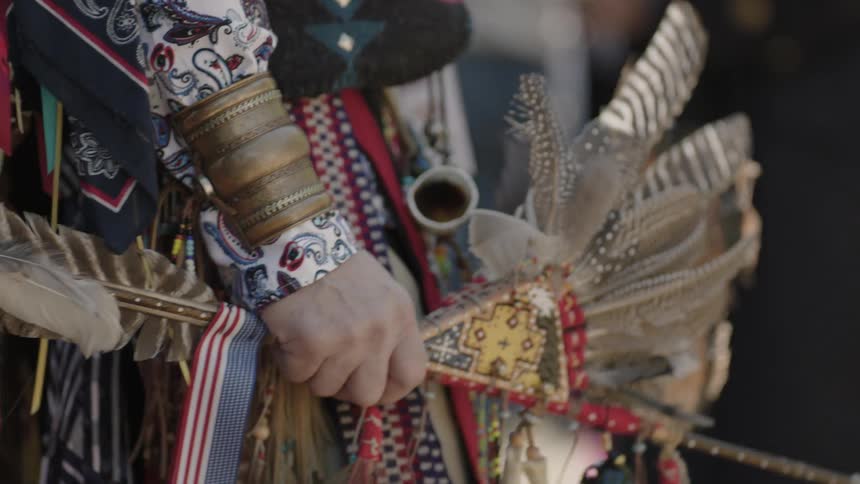 A person wearing a native costume adorned with feathers holds a decorated rod