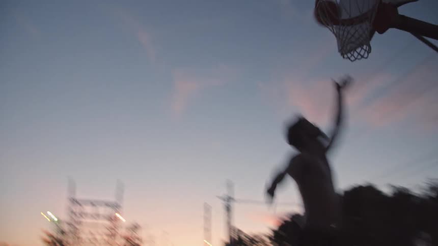 Silhouette of a young man playing basketball by himself on an outdoors court at evening