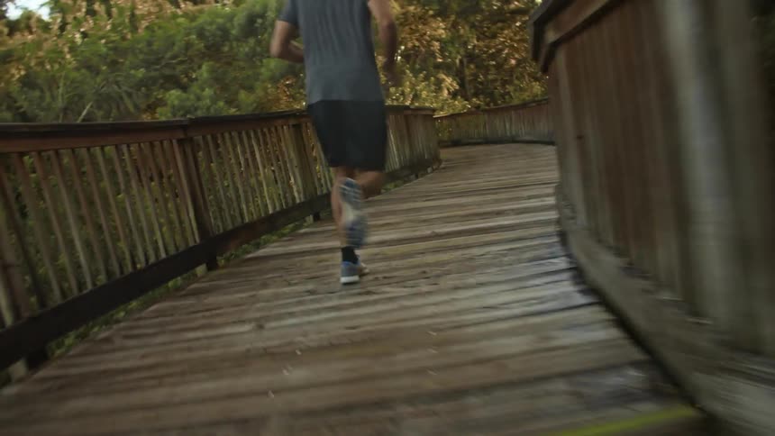 Man with gray t shirt shorts and a pair of sneakers runs across wood bridge in the forest