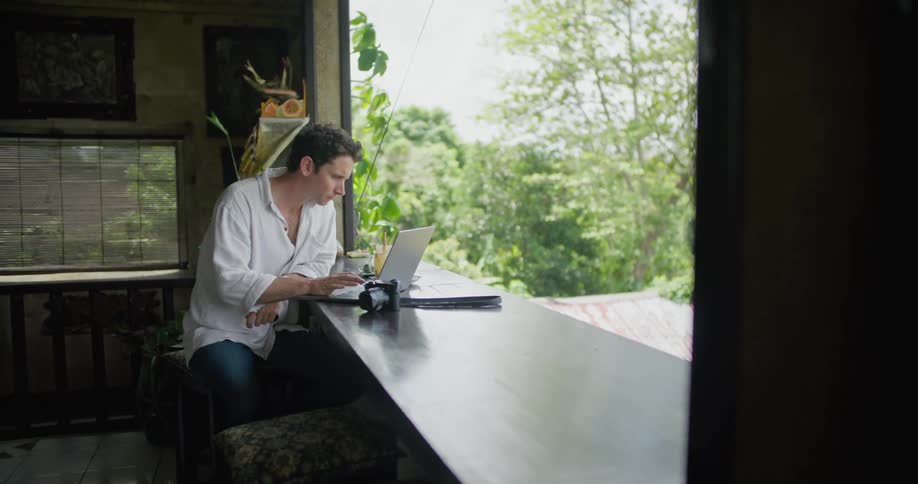 A man is sitting at a counter with a laptop computer as he drinks his coffee and types on the keyboard and looks outside