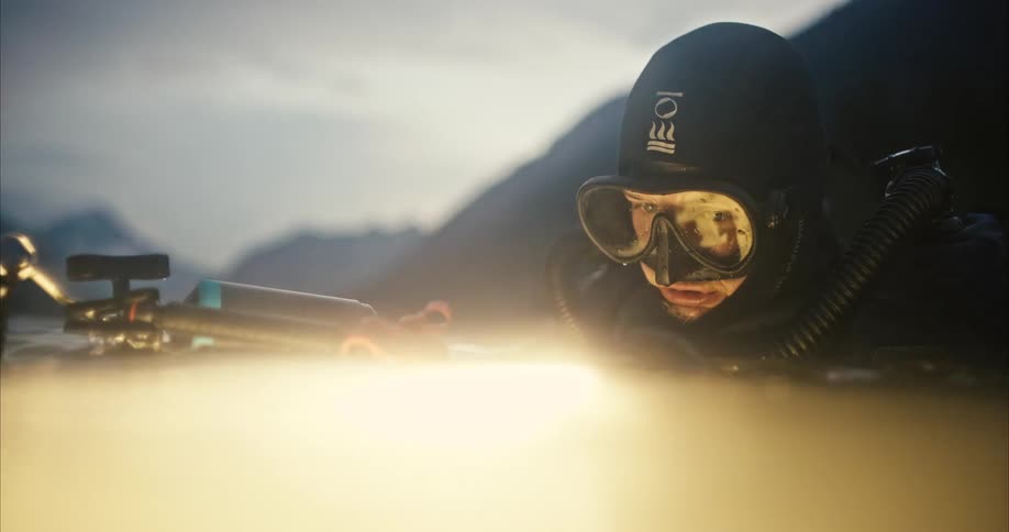 A scuba diver holds an underwater camera near the surface with hills and land behind him