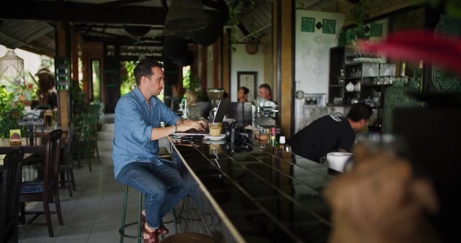 Man sitting at a bar counter using his laptop while the bartender is working in front of him
