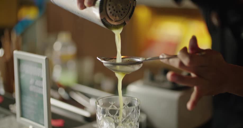 A bartender pours a liquid from a shaker through a strainer and into a drinking glass on a bar counter