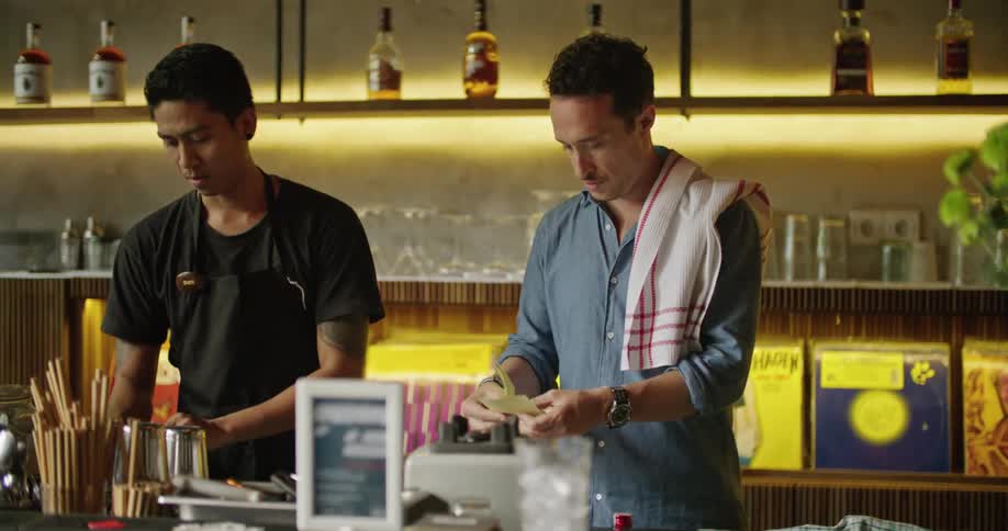 A bartender is standing behind the bar slicing limes as another bartender is talking to him