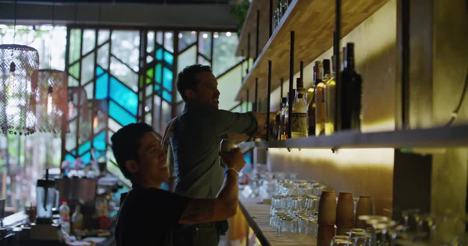 Two bartenders are working together stocking and displaying bottles on the shelf behind the bar