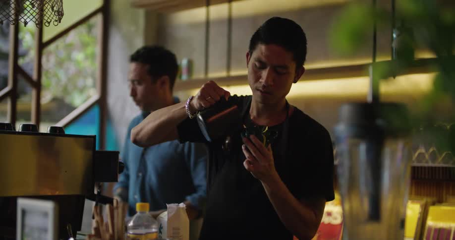 A man pours a coffee in a coffee shop as another man cleans the espresso machine